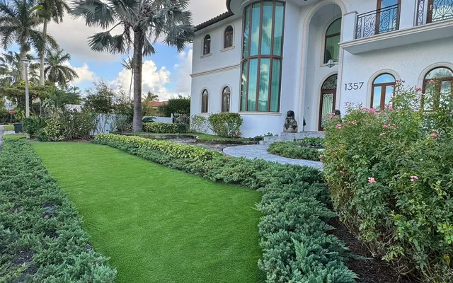 Lush artificial turf side yard bordered by hedges, with a tall palm canopy casting dappled shade across the lawn